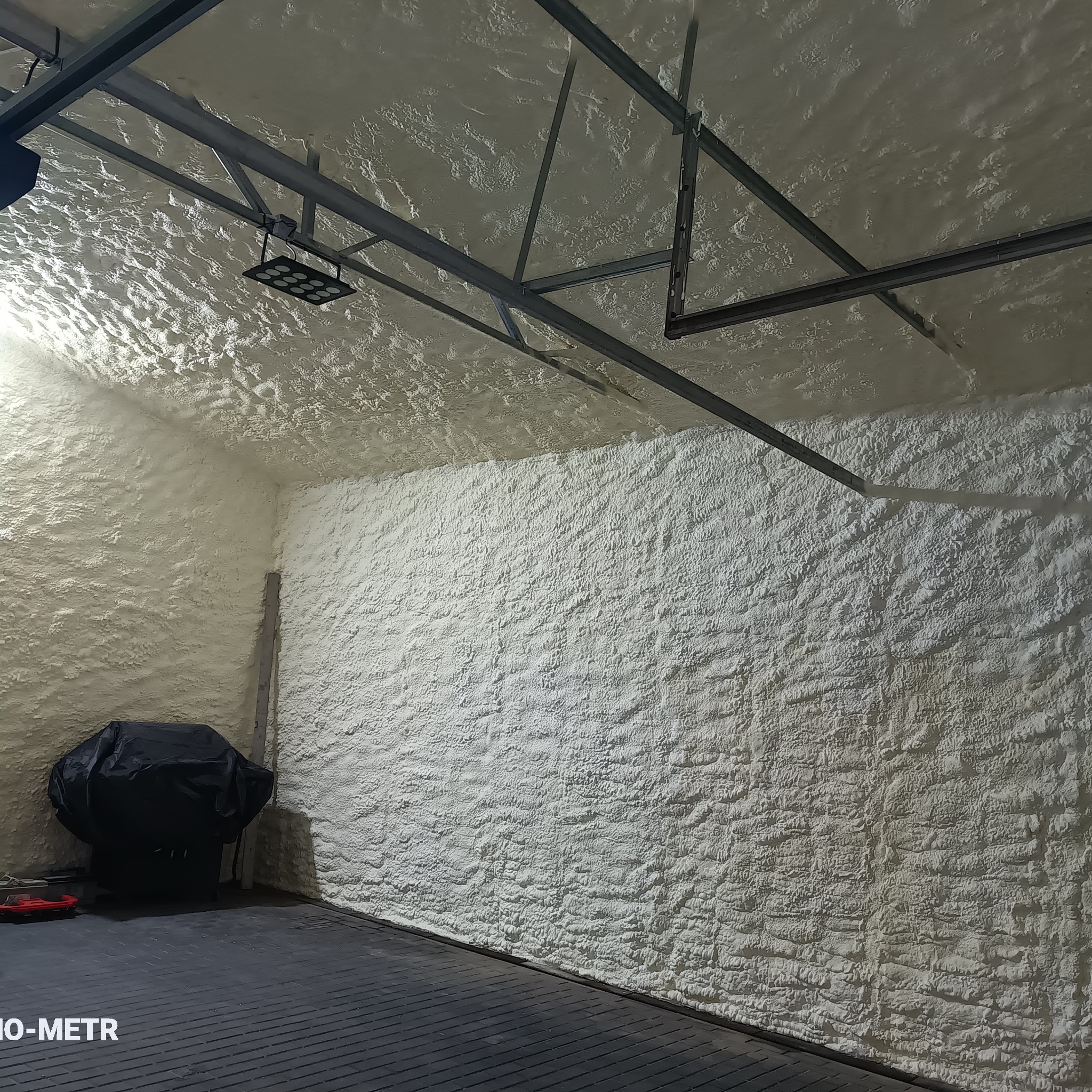 Poddasze po ociepleniu pianką PUR: biała, nierówna powierzchnia izolacji pokrywa ściany i sufit, widoczne elementy konstrukcji dachu i oświetlenie. W rogu stoi grill pod pokrowcem.