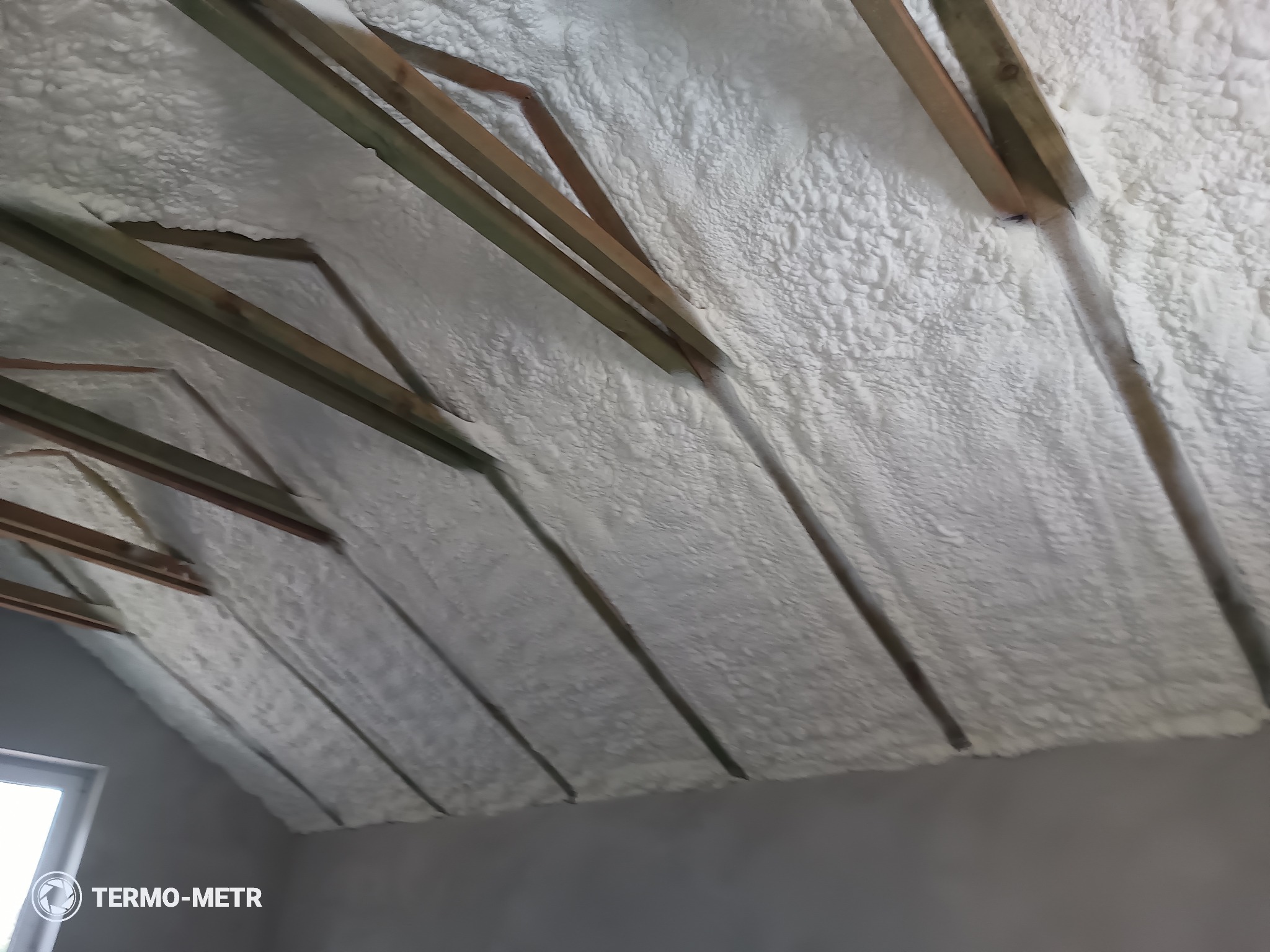 Poddasze z drewnianą konstrukcją pokryte białą pianką izolacyjną. Widoczne krokwie i fragment okna. Tekst TERMO-METR w lewym dolnym rogu.