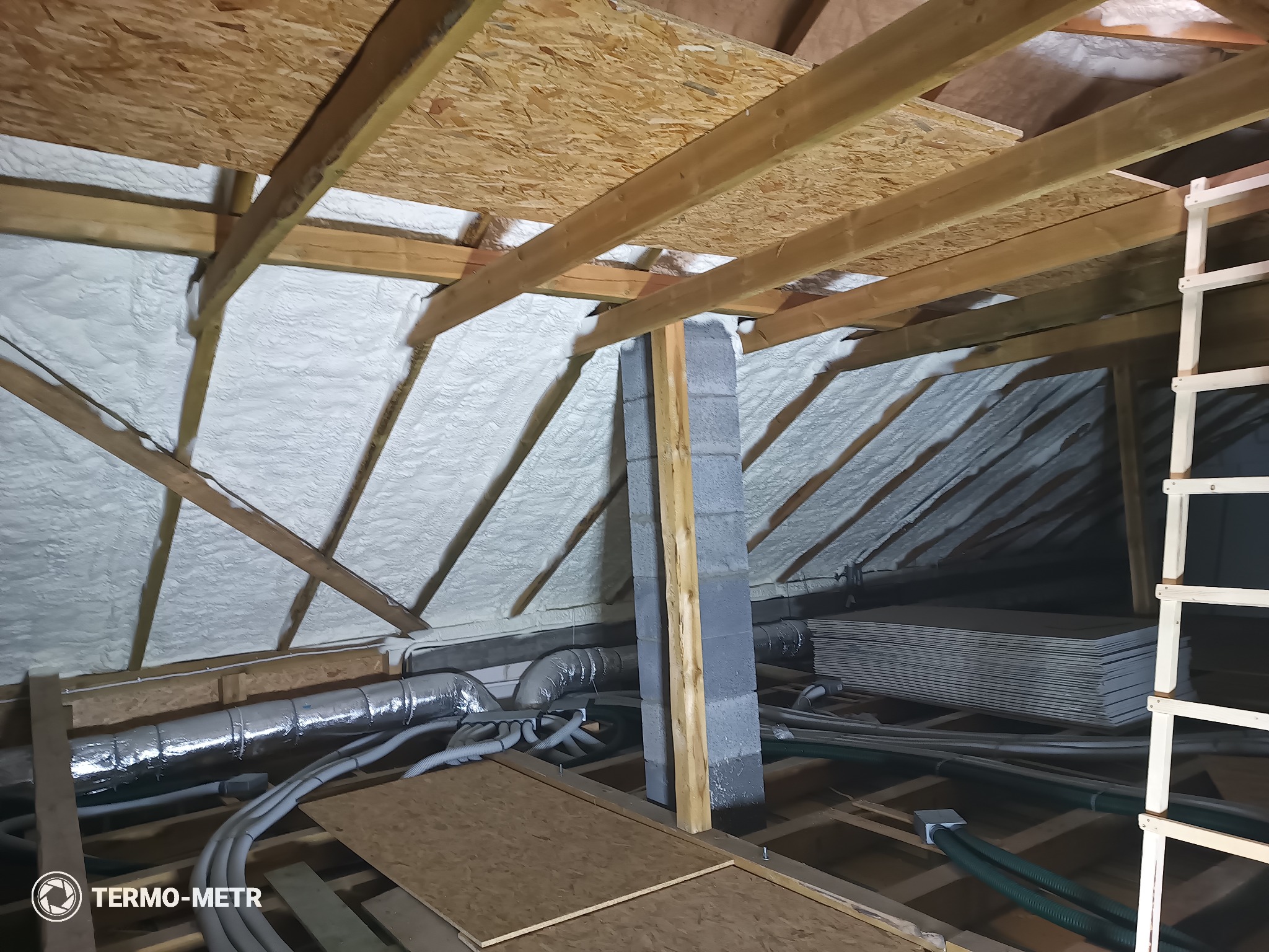 Poddasze z drewnianą konstrukcją i szarą pianką izolacyjną, widoczne przewody wentylacyjne i komin z bloczków. Ujęcie z dołu, z widocznym logo firmy w rogu.