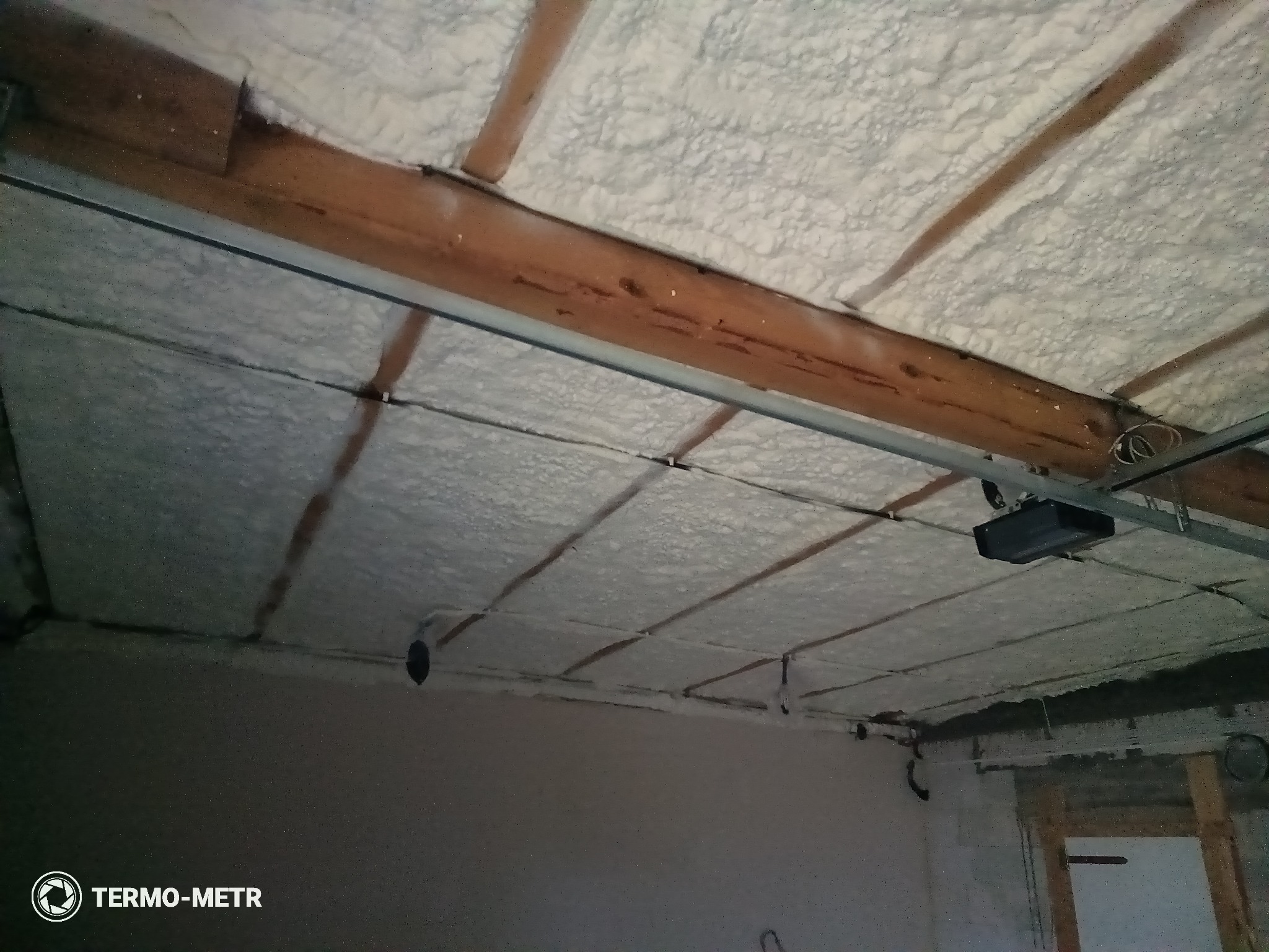 Poddasze z drewnianą konstrukcją i białą pianką izolacyjną, widoczne elementy instalacji. Ocieplenie wykonane natryskowo, równomierna warstwa pianki.