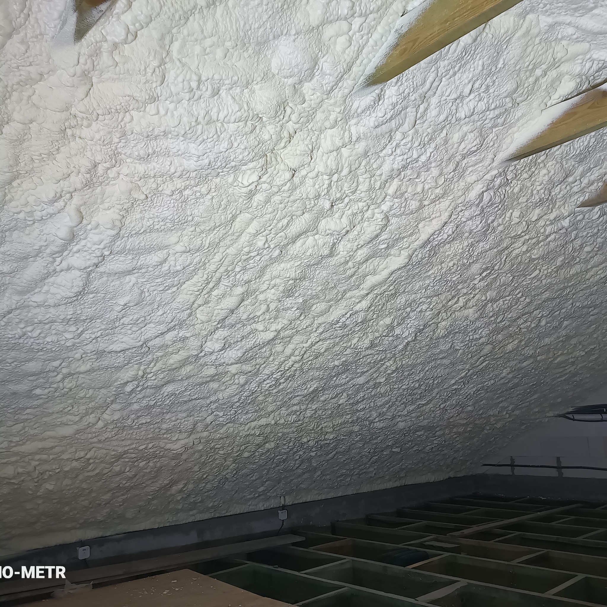 Poddasze z widoczną strukturą natryśniętej, jasnej pianki izolacyjnej, z fragmentami drewnianej konstrukcji dachu. Widoczna podłoga z drewnianych belek.