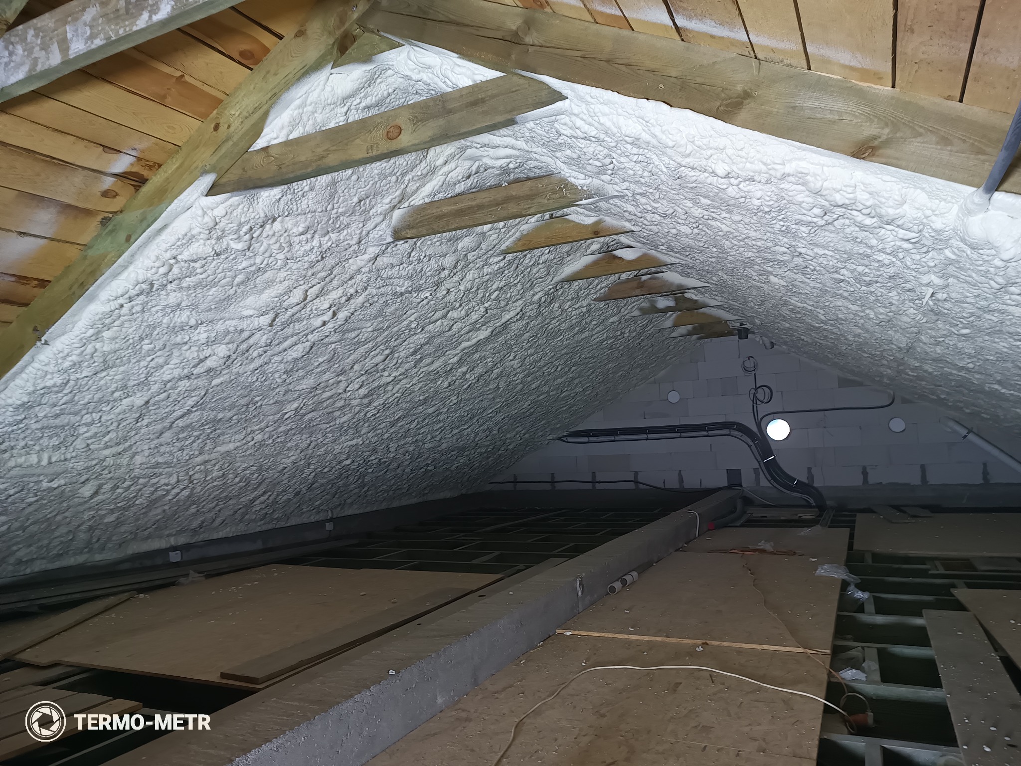 Poddasze z drewnianą konstrukcją pokryte grubą warstwą białej pianki izolacyjnej. Widoczne elementy instalacji elektrycznej i wentylacyjnej. Ujęcie z dołu.