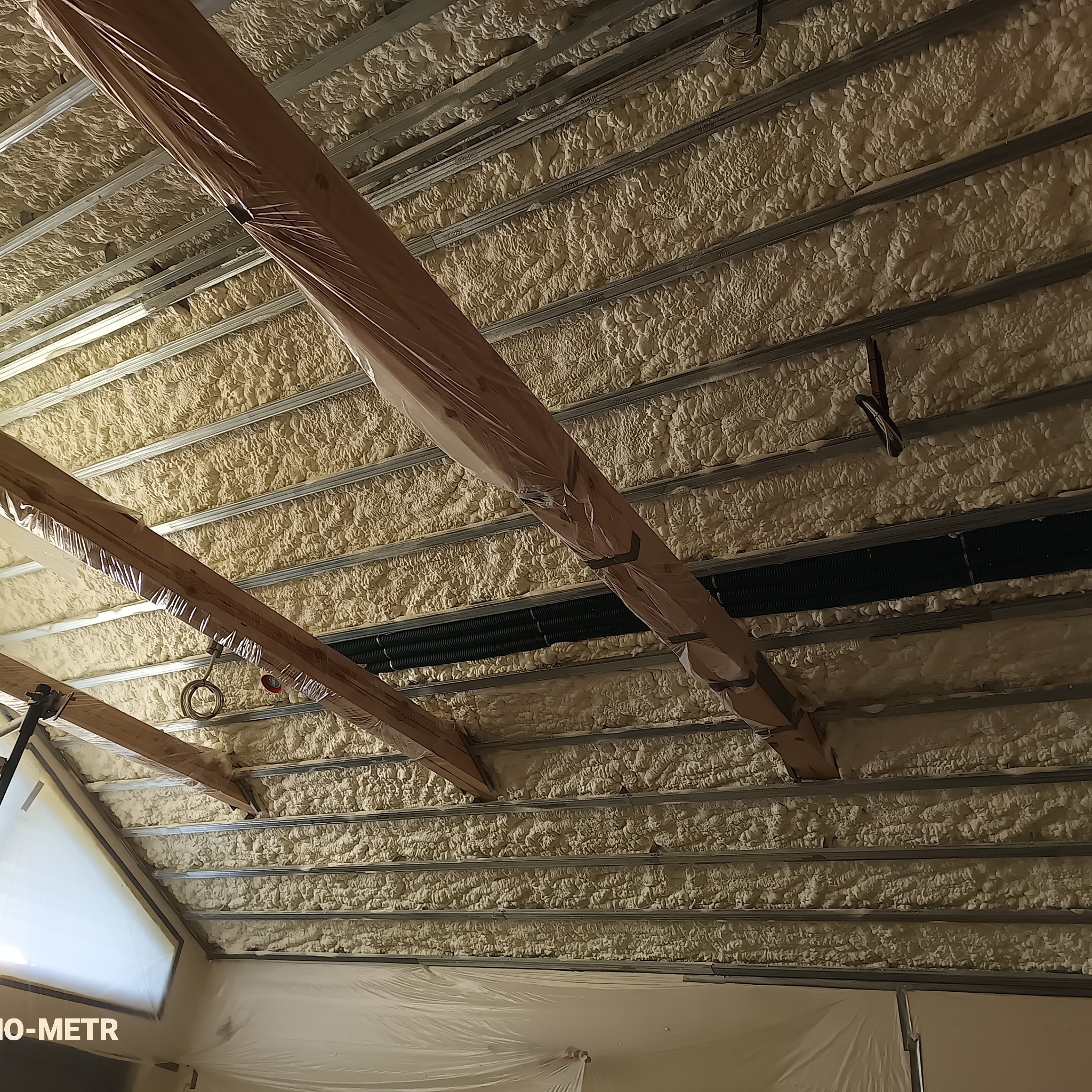 Poddasze ocieplone pianką PUR, widoczne metalowe profile i drewniane belki zabezpieczone folią. Struktura pianki gęsta, równomiernie nałożona. Ujęcie z dołu.