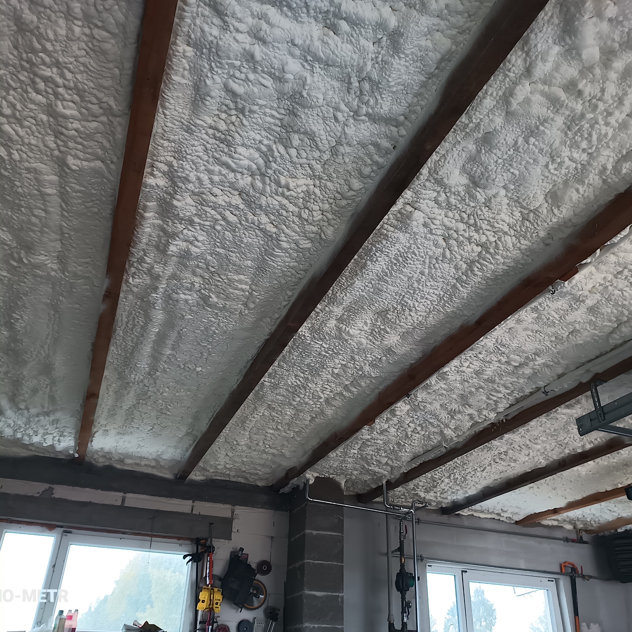 Poddasze ocieplone pianką PUR, widoczne drewniane belki konstrukcyjne. Biała, nierówna powierzchnia pianki pokrywa całą przestrzeń między belkami. Widok z dołu.