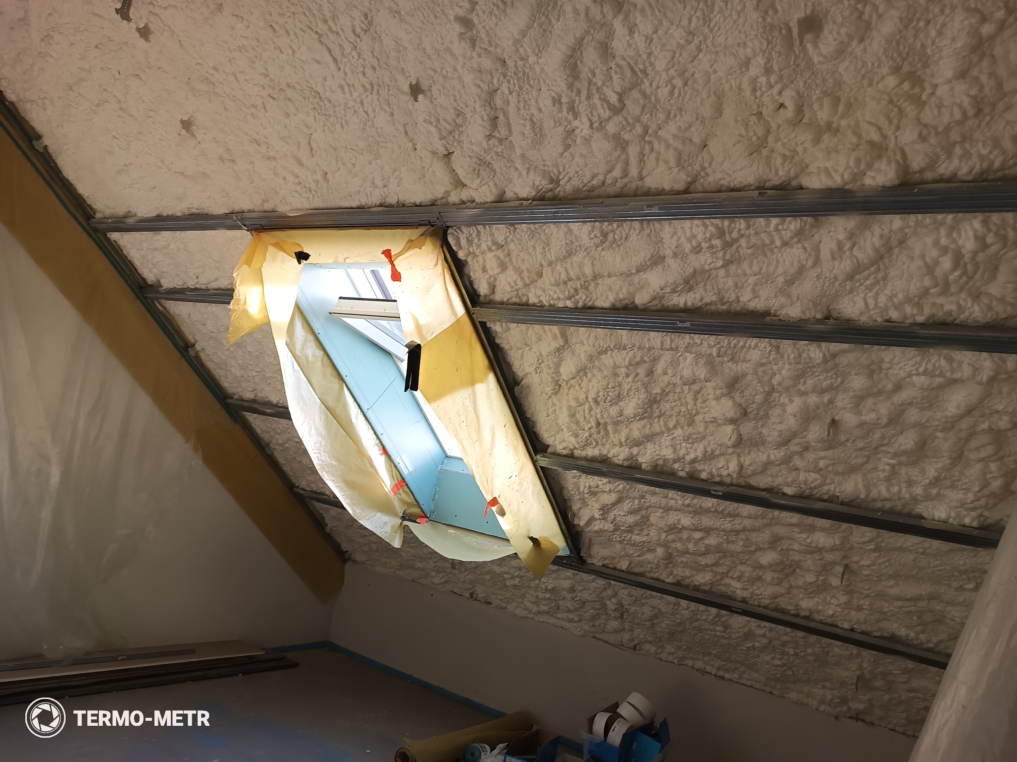Poddasze po ociepleniu pianką PUR w Białej Podlaskiej. Widoczne metalowe profile i okno dachowe zabezpieczone folią. Izolacja natryskowa, równomierna struktura piany.