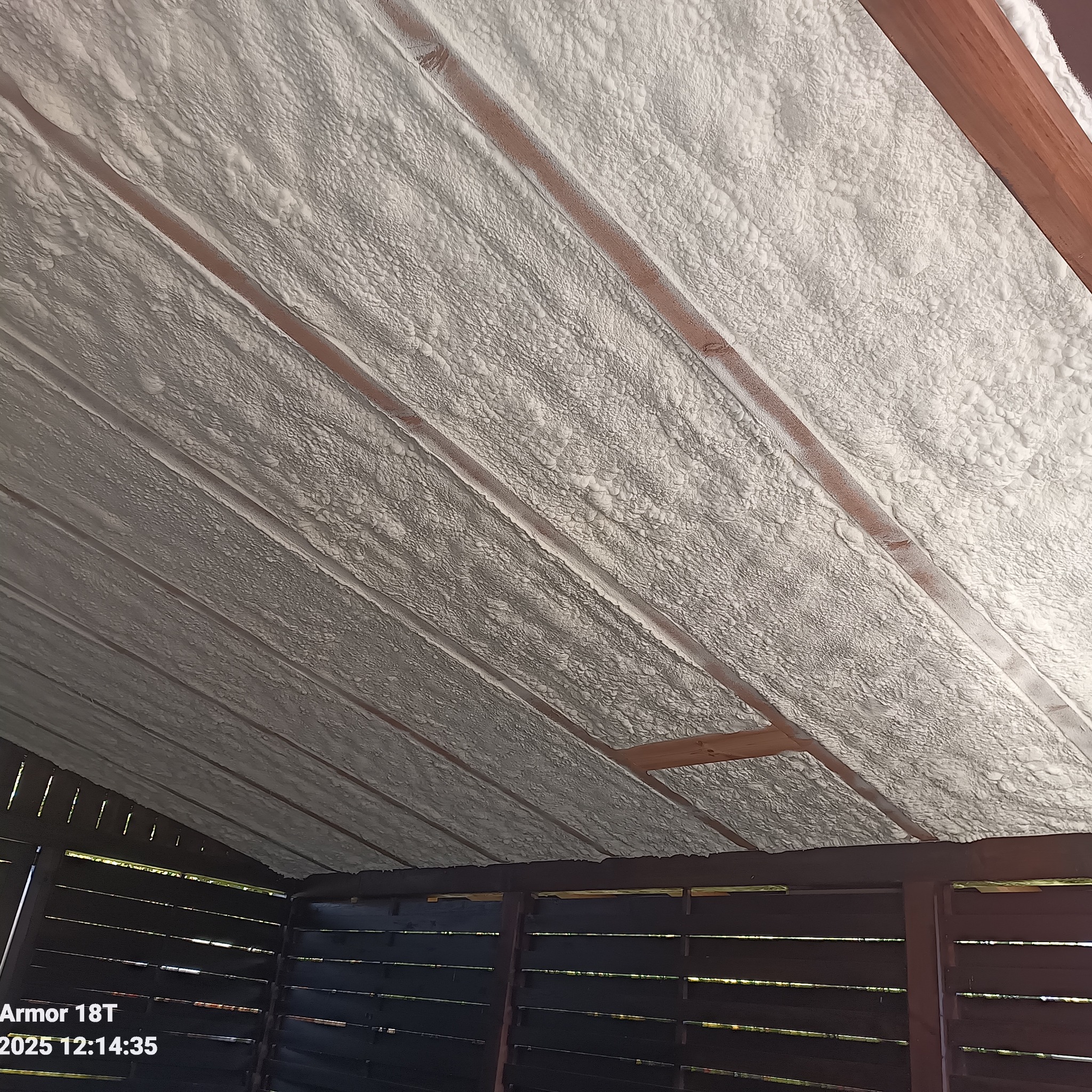 Poddasze z widoczną strukturą pianki izolacyjnej, aplikowaną między krokwiami. Widoczne drewniane elementy konstrukcji dachu. Ujęcie z dołu, pod kątem.