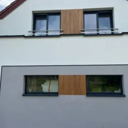 Elewacja budynku po remoncie z oknami, częściowo zasłoniętymi płytami imitującymi drewno; w jednym oknie widoczne płyty ociepleniowe.