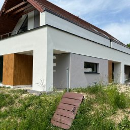 USŁUGI REMONTOWE RAFAŁ HABAS - Nowoczesny dom jednorodzinny z białymi ścianami i drewnianymi panelami, widoczne ślady prac wykończeniowych na trawniku, w tle worki z odpadami budowlanymi.