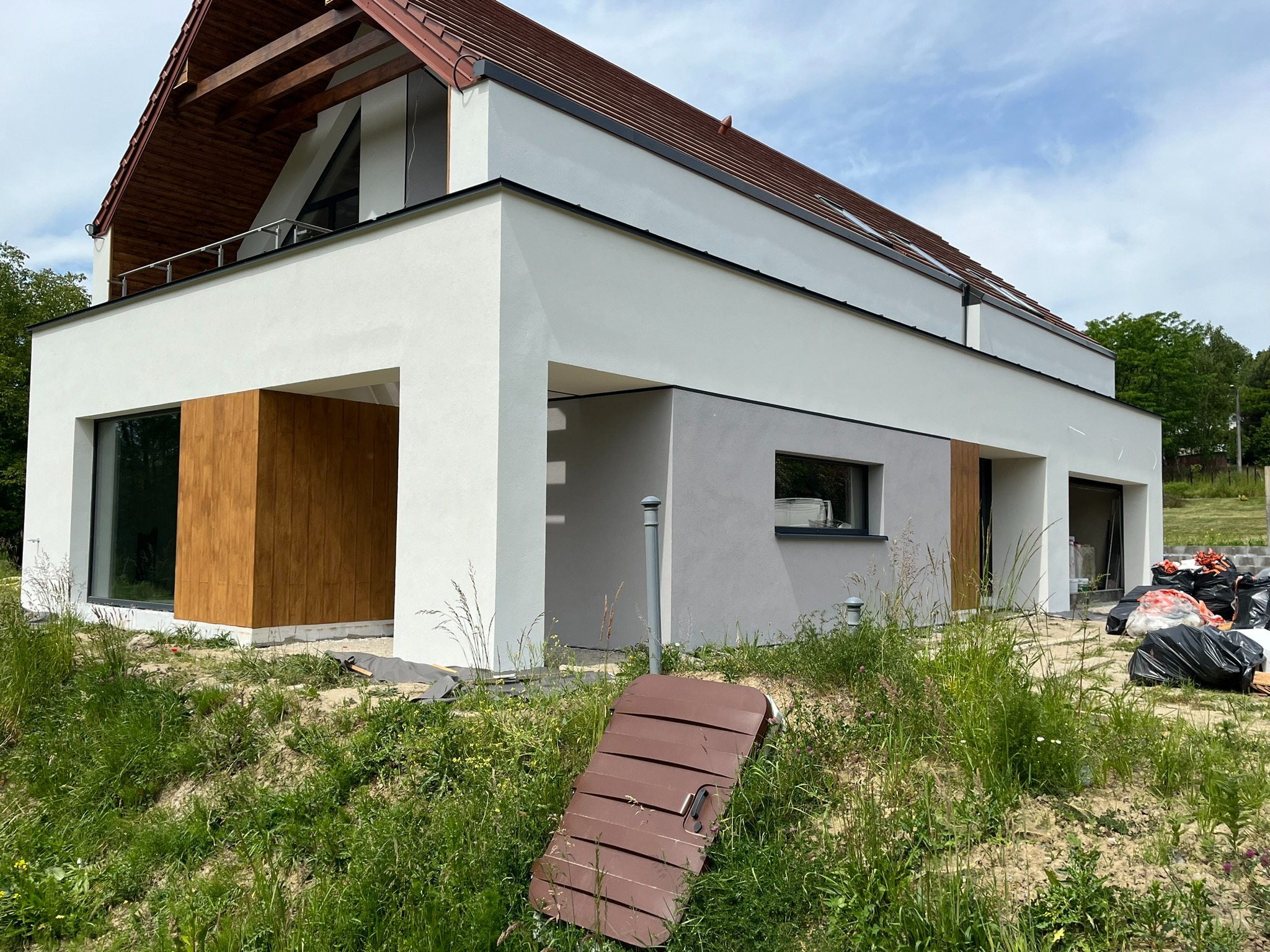 Nowoczesny dom jednorodzinny z białymi ścianami i drewnianymi panelami, widoczne ślady prac wykończeniowych na trawniku, w tle worki z odpadami budowlanymi.