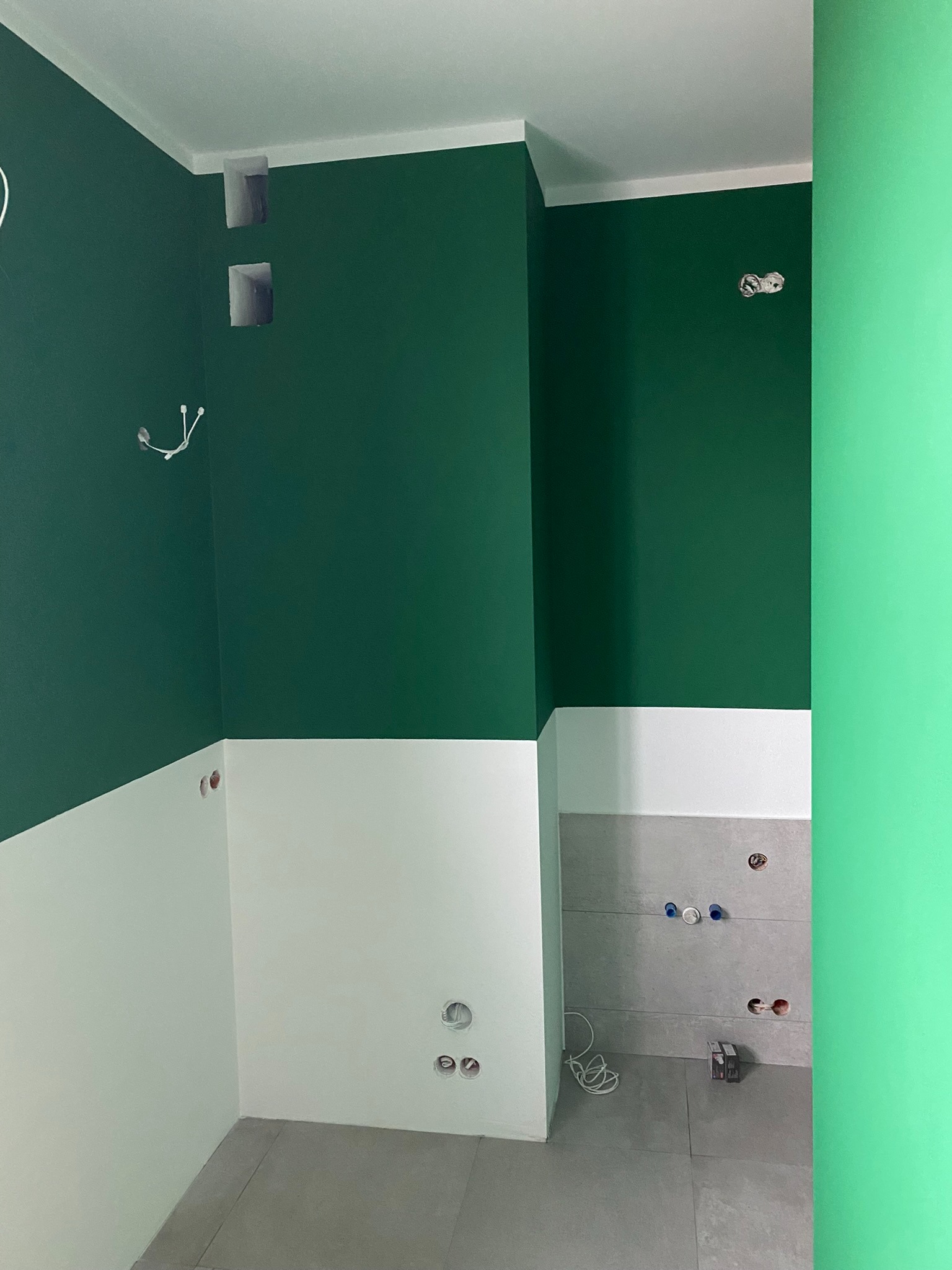 Ściana w trakcie wykończenia: górna część pomalowana na ciemnozielono, dolna na biało, widoczne puszki elektryczne i przewody, szara glazura w tle.