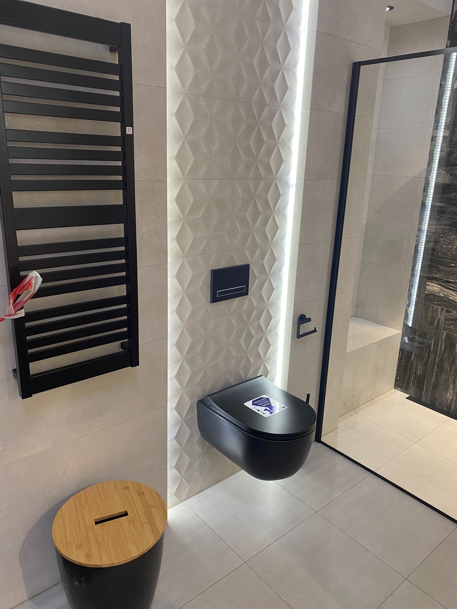 Nowoczesna łazienka z czarną wiszącą toaletą, grzejnikiem drabinkowym i kabiną prysznicową z czarnymi ramami, oświetlona pionowym paskiem LED.