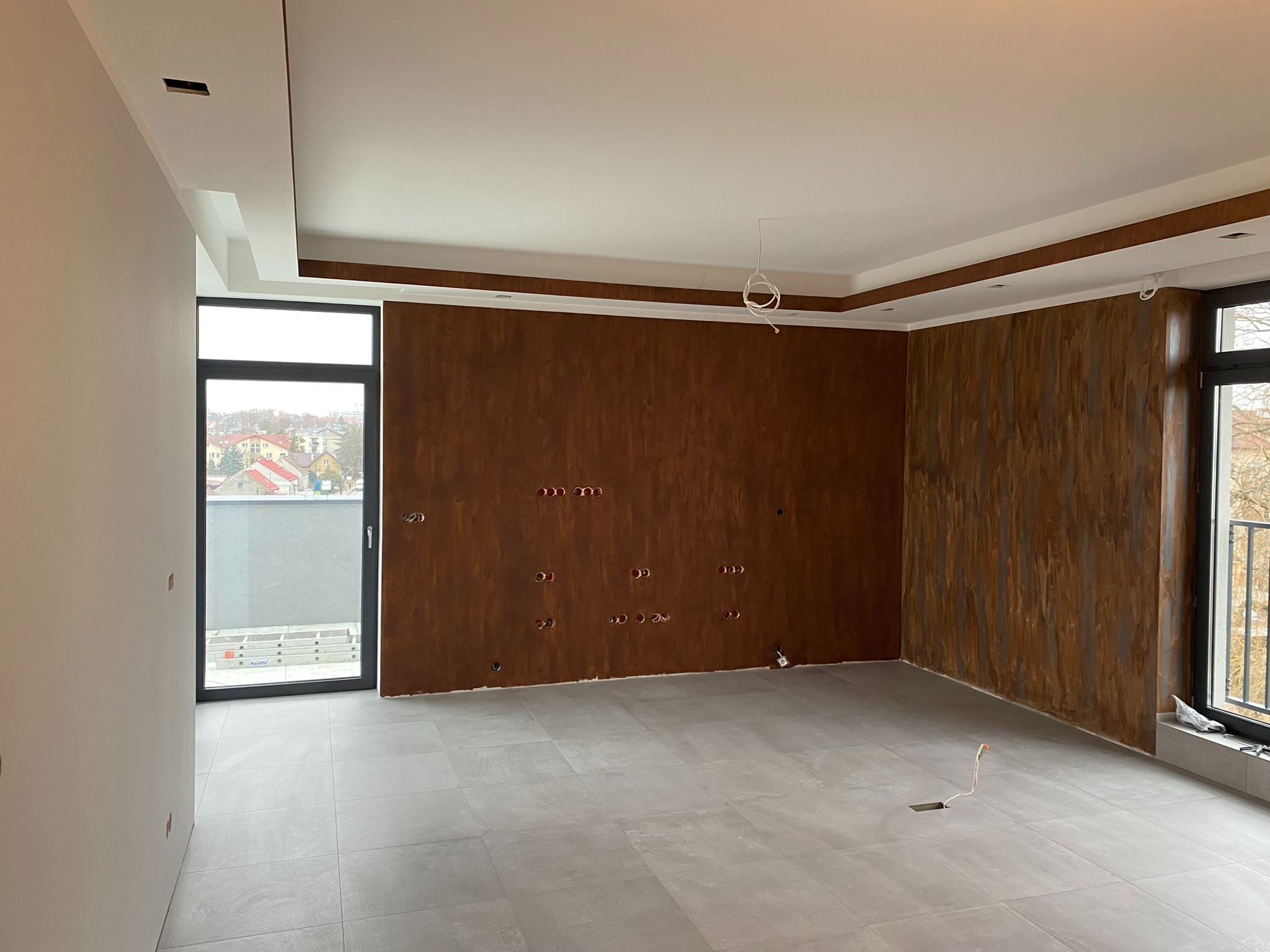 Pusty pokój po malowaniu ścian na brązowo z widocznymi otworami na gniazdka, szara podłoga z płytek, okno z widokiem na miasto, sufit z dekoracyjnymi listwami.