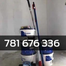 Stos wiader z farbą Sigma, wałki malarskie z teleskopowymi uchwytami oraz kuweta na farbę w narożniku pomalowanego na szaro pokoju.