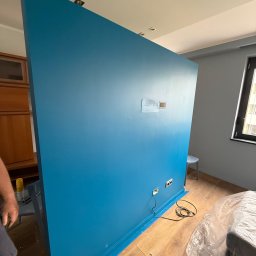 TDM House - Świeżo pomalowana na turkusowo ściana z gniazdkami i puszką elektryczną, widoczna w trakcie remontu wnętrza. Częściowo zakryte meble folią ochronną.