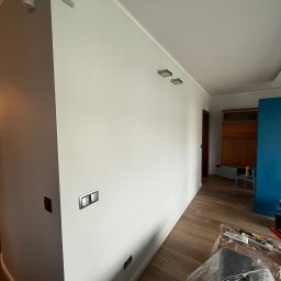 TDM House - Jasne, świeżo pomalowane wnętrze z drewnianą podłogą. Widoczne detale: czujniki na ścianie, nowoczesne włączniki i zabezpieczone meble. Neutralna kolorystyka.