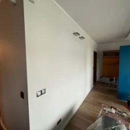 Jasne, świeżo pomalowane wnętrze z drewnianą podłogą. Widoczne detale: czujniki na ścianie, nowoczesne włączniki i zabezpieczone meble. Neutralna kolorystyka.