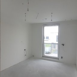 TDM House - Pokój po szpachlowaniu i gruntowaniu, przygotowany do malowania. Widoczne gładkie ściany, okno z widokiem na budynek i wystające kable oświetleniowe.