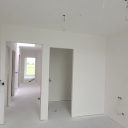 TDM House - Jasne, przestronne wnętrze po szpachlowaniu i malowaniu na biało. Widoczne gładkie ściany, sufit z wystającymi przewodami elektrycznymi i otwarte przejścia do innych pomieszczeń.