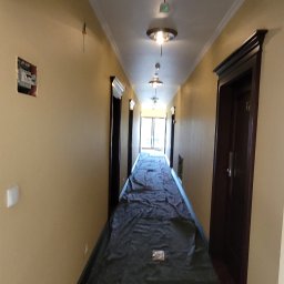 TDM House - Korytarz w budynku w trakcie prac przygotowawczych do malowania. Podłoga zabezpieczona folią ochronną. Widoczne ściany w odcieniach beżu, lampy sufitowe.