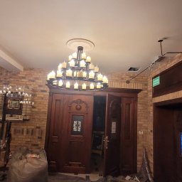 TDM House - Przygotowanie do malowania: zabezpieczona podłoga, wiszący żyrandol i ceglana ściana w tle. Widoczny worek z odpadami i otwarte, drewniane drzwi.