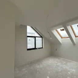 Wnętrze poddasza po gładziowaniu ścian i sufitu, z oknami dachowymi i trójkątnym oknem. Na podłodze widoczne ślady po pracach wykończeniowych.