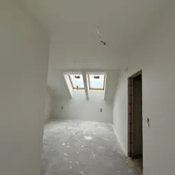 Wnętrze poddasza po remoncie, świeżo pomalowane na biało, z dwoma oknami dachowymi i widocznymi gniazdkami elektrycznymi. Podłoga zabezpieczona folią malarską.