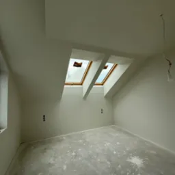 Jasne poddasze z dwoma oknami dachowymi i gładkimi ścianami po szpachlowaniu, przygotowane do malowania. Widoczne puszki elektryczne i surowa podłoga.