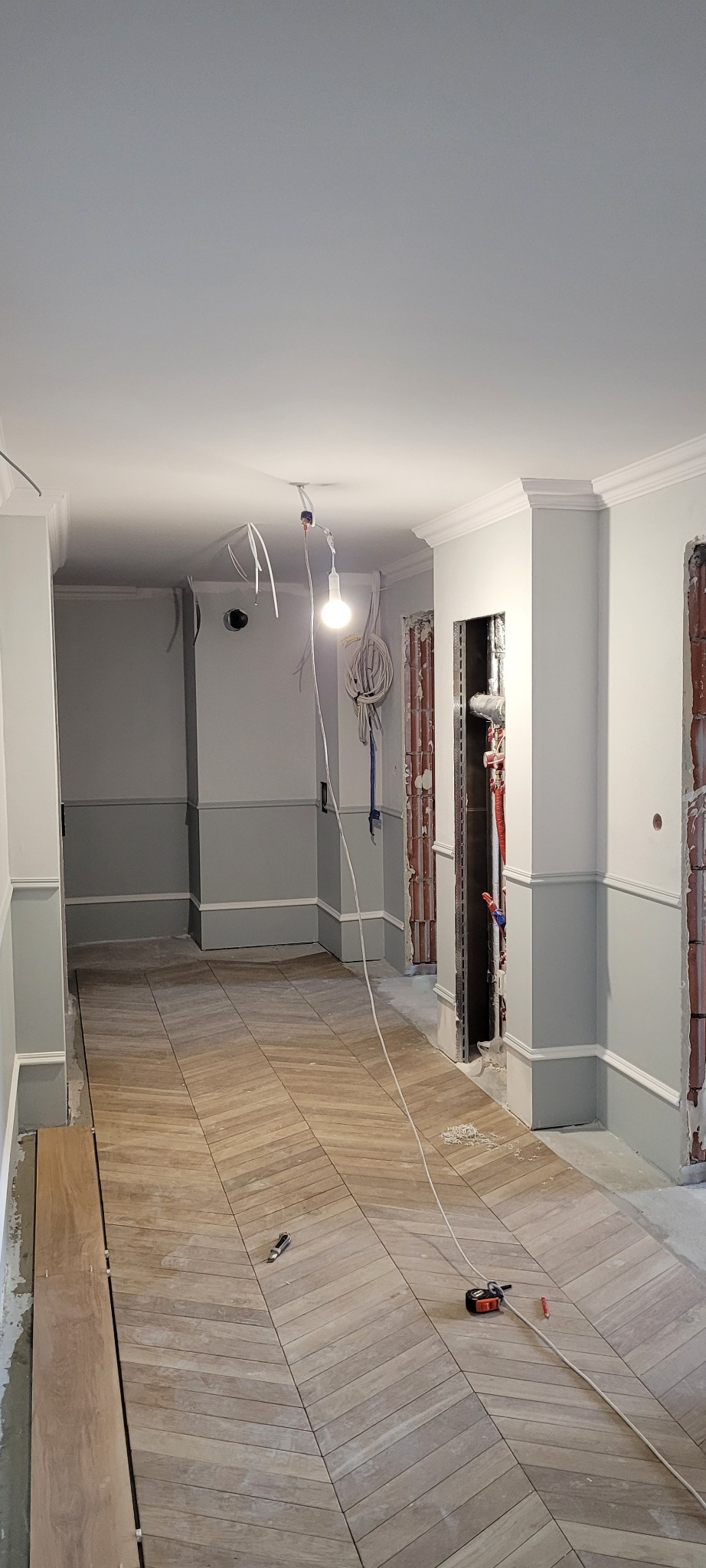 Remont korytarza we Wrocławiu w trakcie prac: położona podłoga z płytek imitujących drewno ułożonych we wzór jodełki, pomalowane ściany do połowy na szaro, widoczne narzędzia i kable elektryczne.