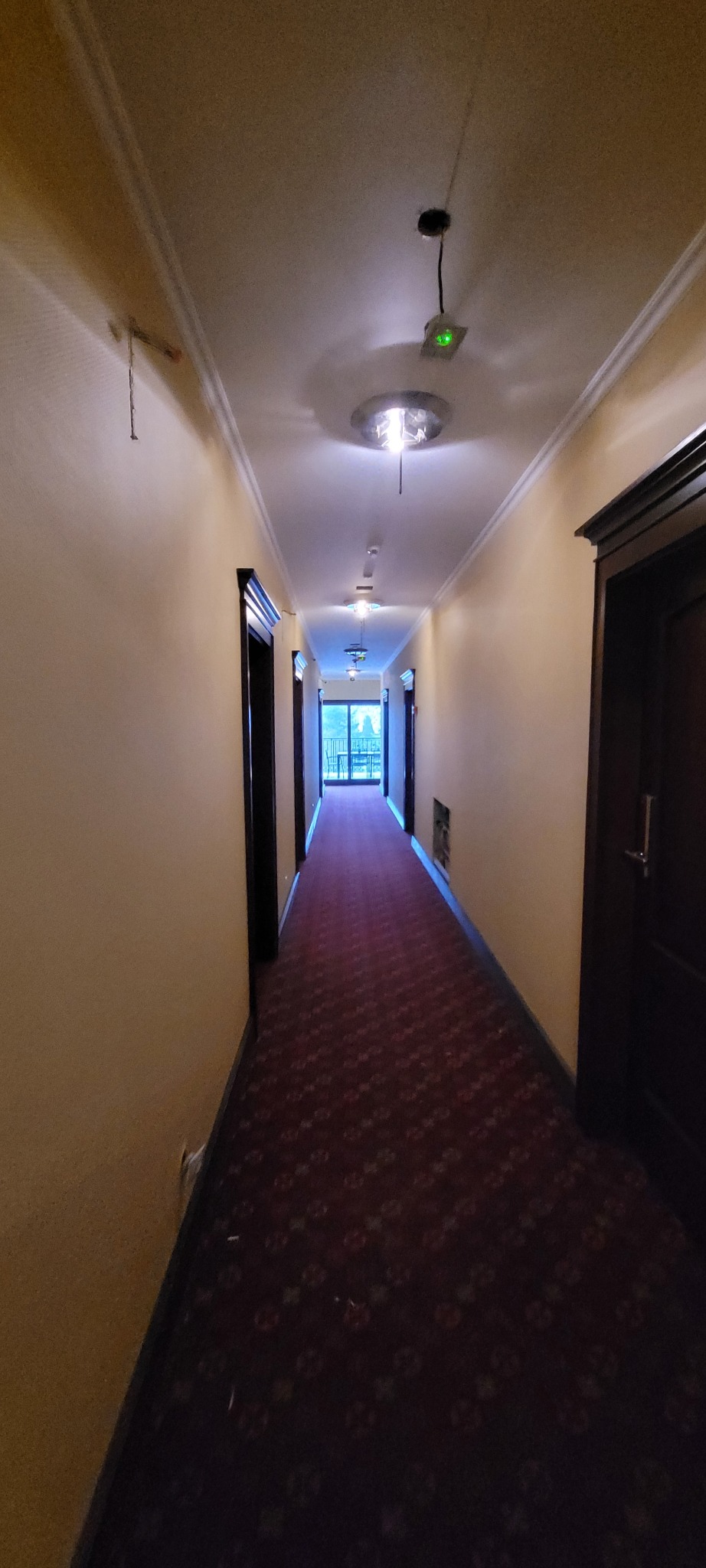 Długi korytarz hotelowy z czerwonym dywanem w kwiaty, kremowymi ścianami, ciemnymi drzwiami i oświetleniem sufitowym, widok na balkon.