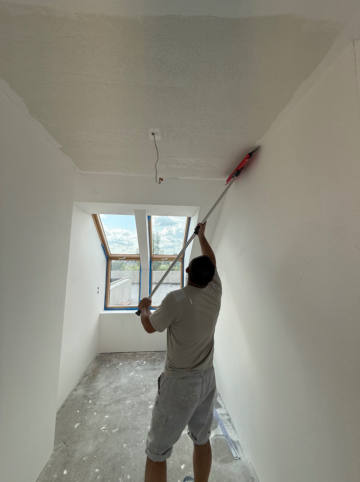 Prace wykończeniowe: mężczyzna szpachluje ścianę w jasnym pokoju z oknem dachowym, używając narzędzia na długim kiju. Widoczny fragment sufitu i podłogi.