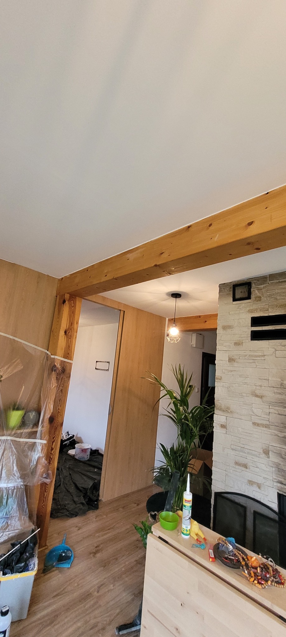 Remont wnętrza: widok na sufit i ścianę przed malowaniem, z drewnianą belką i osłoniętą folią ścianą. W tle widoczne narzędzia i przygotowania do prac malarskich.