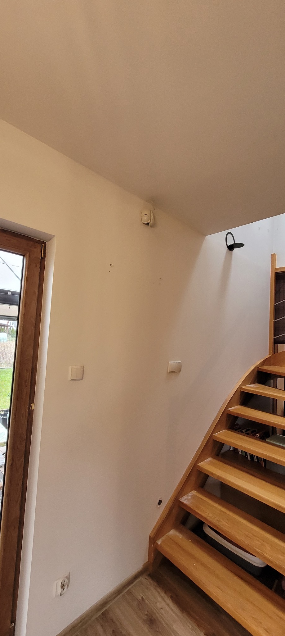 Jasne wnętrze domu po odświeżeniu: kremowe ściany, drewniane schody i rama okienna. Widoczne gniazdka, włączniki i czujnik dymu na ścianie. Minimalistyczny wystrój.