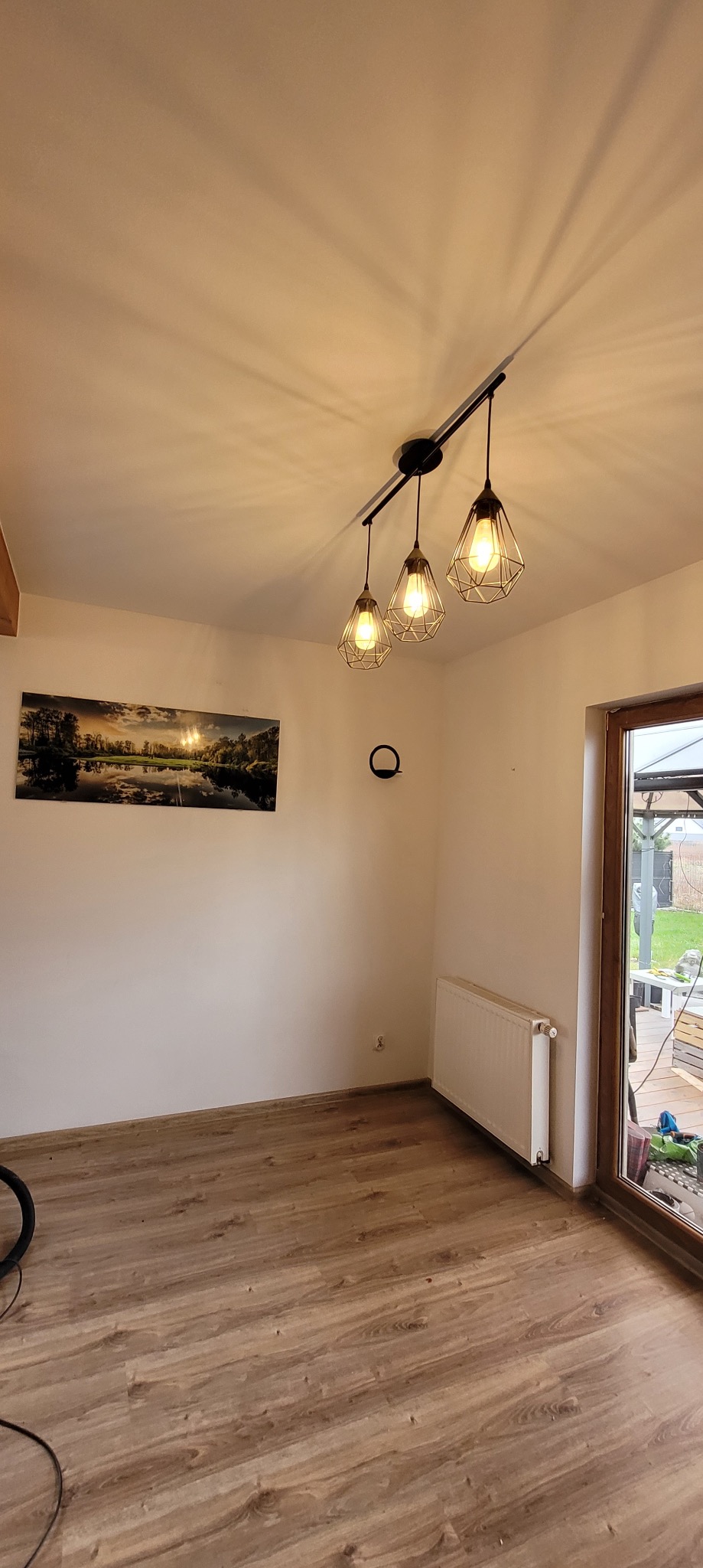 Wnętrze pokoju po remoncie z drewnianą podłogą, białymi ścianami, obrazem i nowoczesną lampą sufitową. Widoczny także grzejnik i okno z widokiem na zewnątrz.