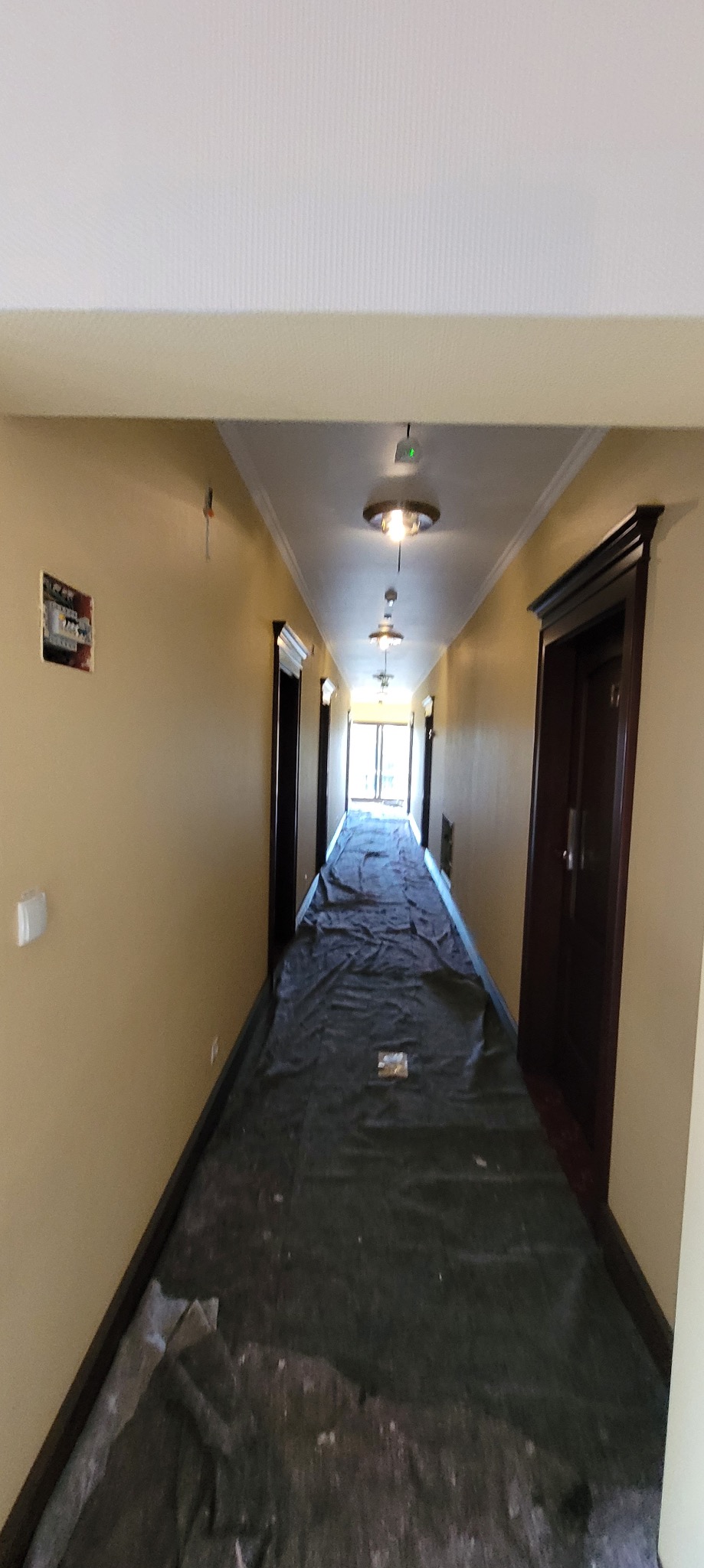 Korytarz w budynku w trakcie prac przygotowawczych do malowania. Podłoga zabezpieczona folią ochronną. Widoczne ściany w odcieniach beżu, lampy sufitowe.