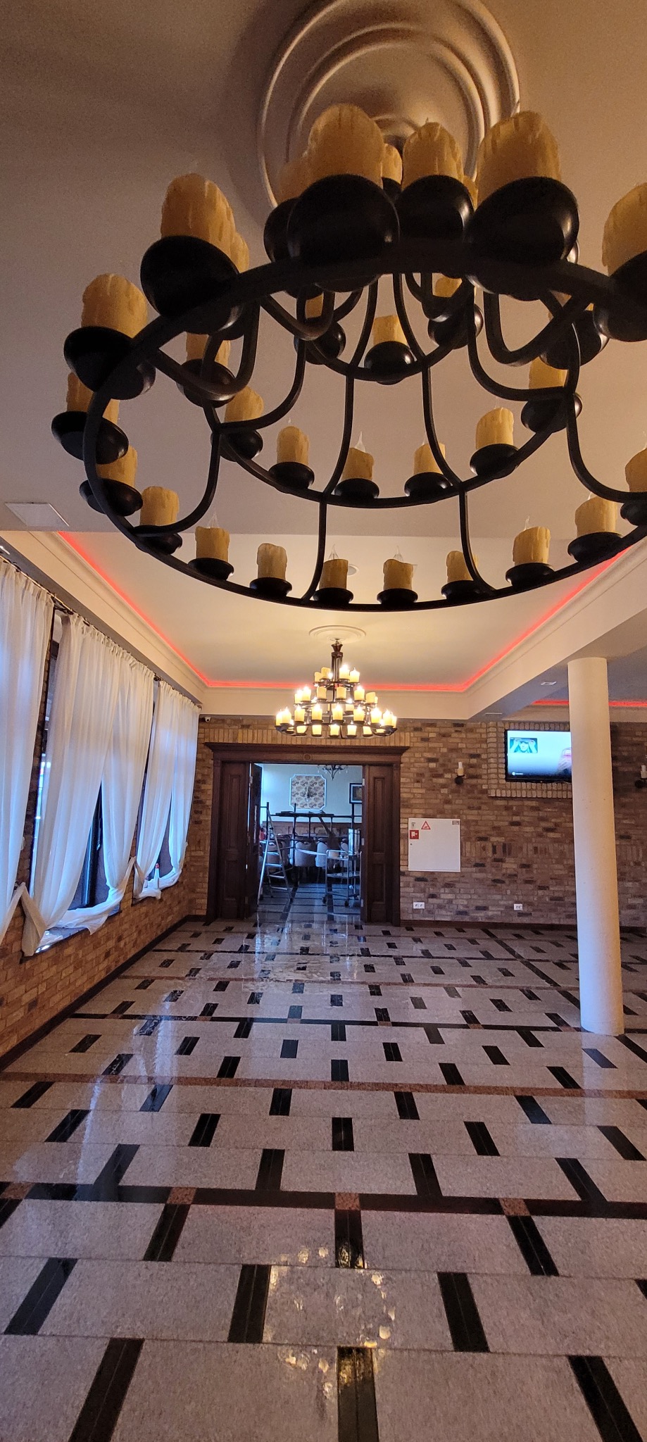 Eleganckie wnętrze z żyrandolami świecznikowymi i marmurową posadzką. Widok na salę bankietową przez otwarte, drewniane drzwi. Czerwone oświetlenie sufitowe.