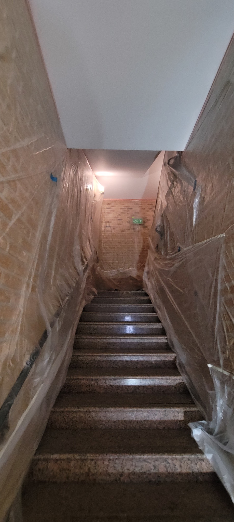Klatka schodowa zabezpieczona folią malarską przed pracami wykończeniowymi. Widoczne schody z lastryko i ceglane ściany. Oświetlenie sufitowe.