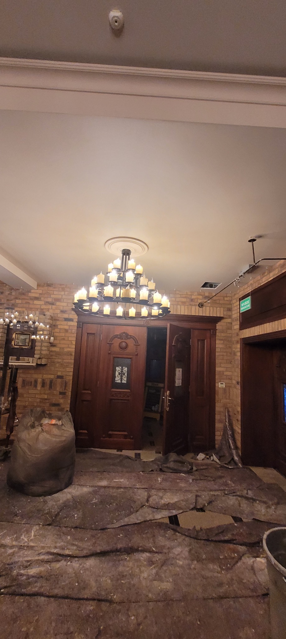 Przygotowanie do malowania: zabezpieczona podłoga, wiszący żyrandol i ceglana ściana w tle. Widoczny worek z odpadami i otwarte, drewniane drzwi.