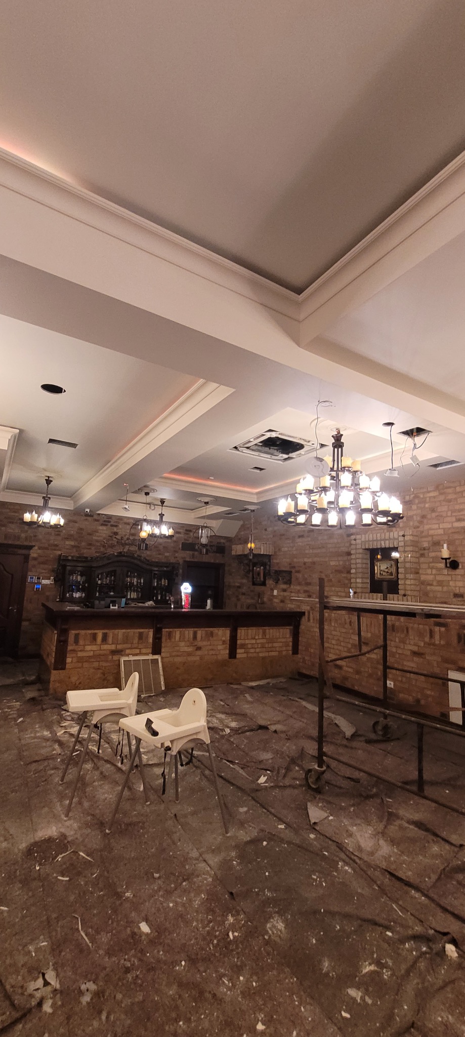 Wnętrze lokalu w trakcie remontu: widoczne odsłonięte instalacje sufitowe, ceglana ściana baru, zabezpieczona podłoga i krzesełka dziecięce. Oświetlenie częściowo zdemontowane.