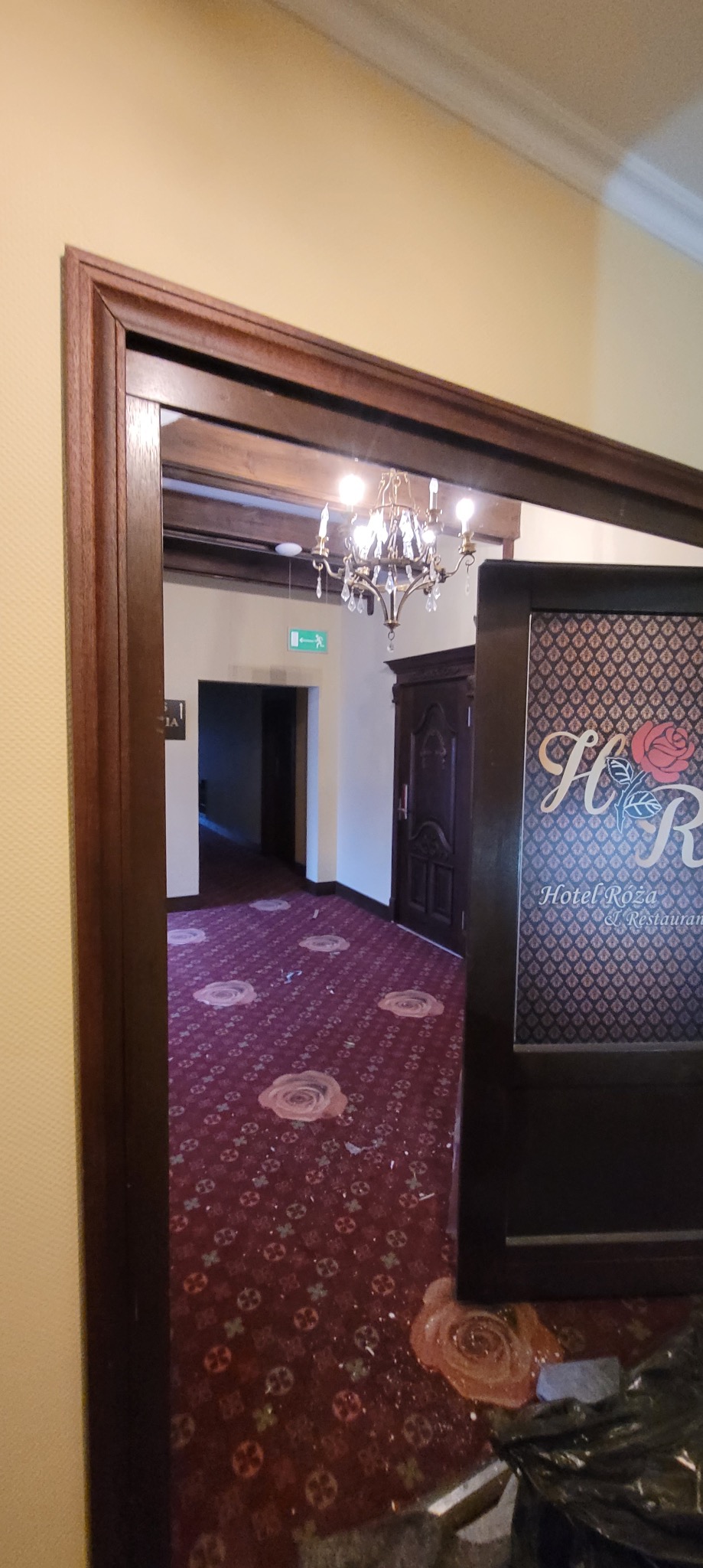 Wnętrze hotelu/restauracji widziane przez futrynę drzwi w trakcie remontu. Widoczny fragment dywanu z wzorem róży i ślady po pracach remontowych.