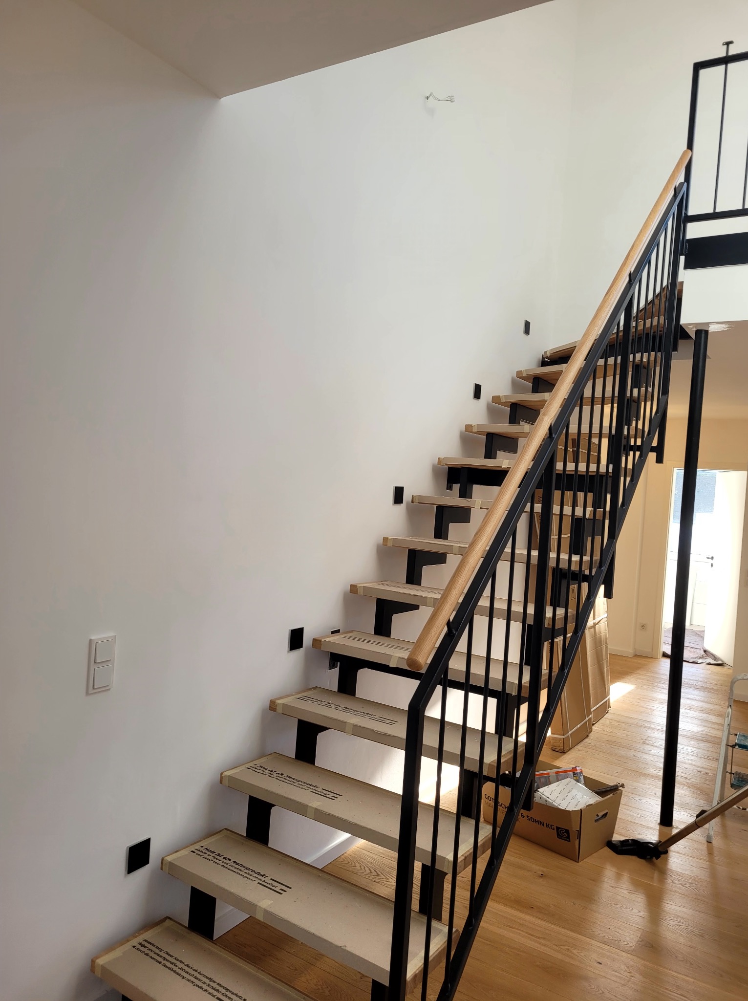 Nowoczesne schody z drewnianymi stopniami i czarnymi metalowymi elementami, zabezpieczone kartonem, na tle świeżo pomalowanej, białej ściany w jasnym wnętrzu.
