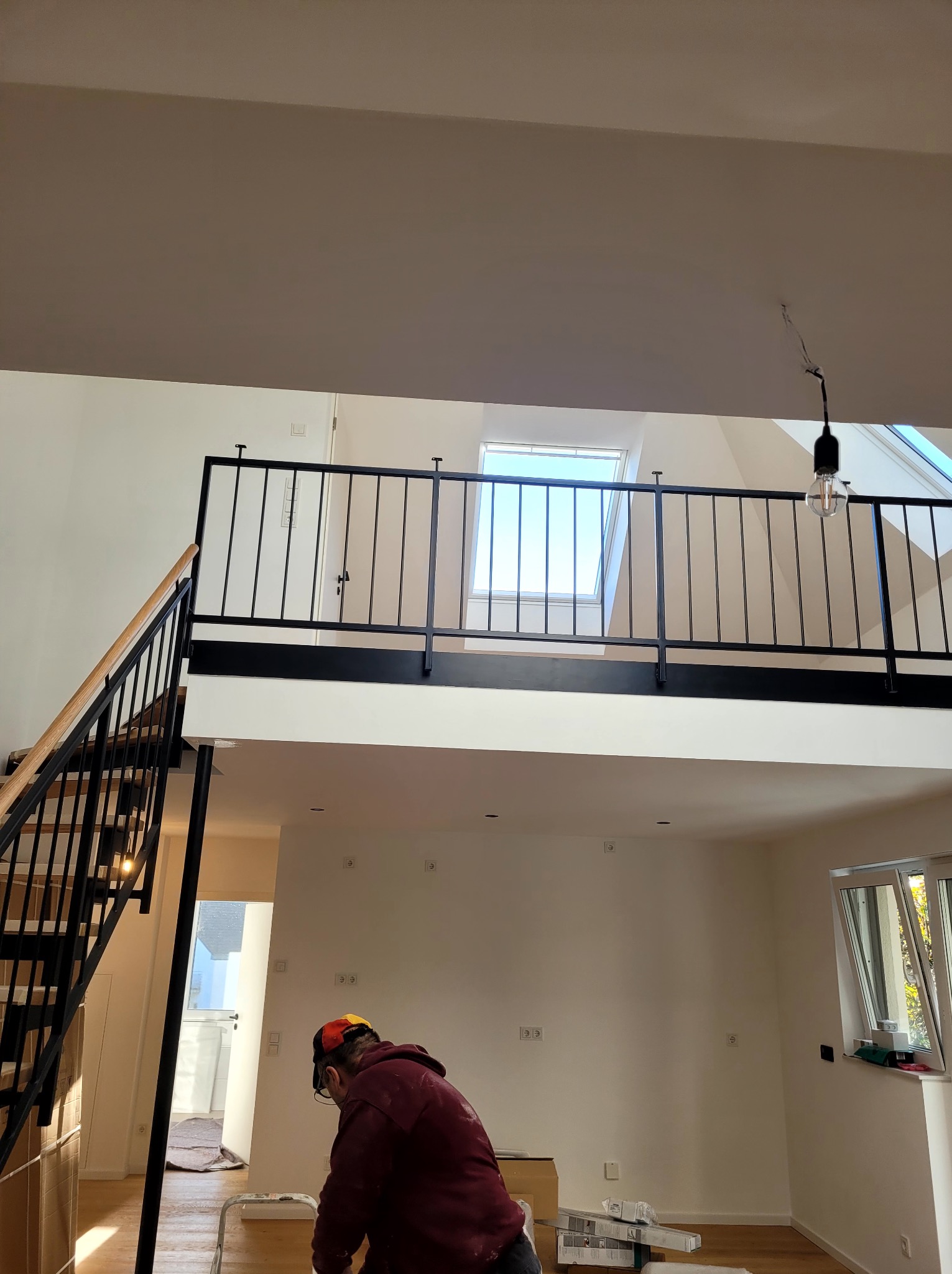 Praca w dwupoziomowym wnętrzu: mężczyzna przygotowuje ściany, widoczne schody z czarną balustradą, okno dachowe i minimalistyczna lampa. Ściany są świeżo pomalowane na biało.