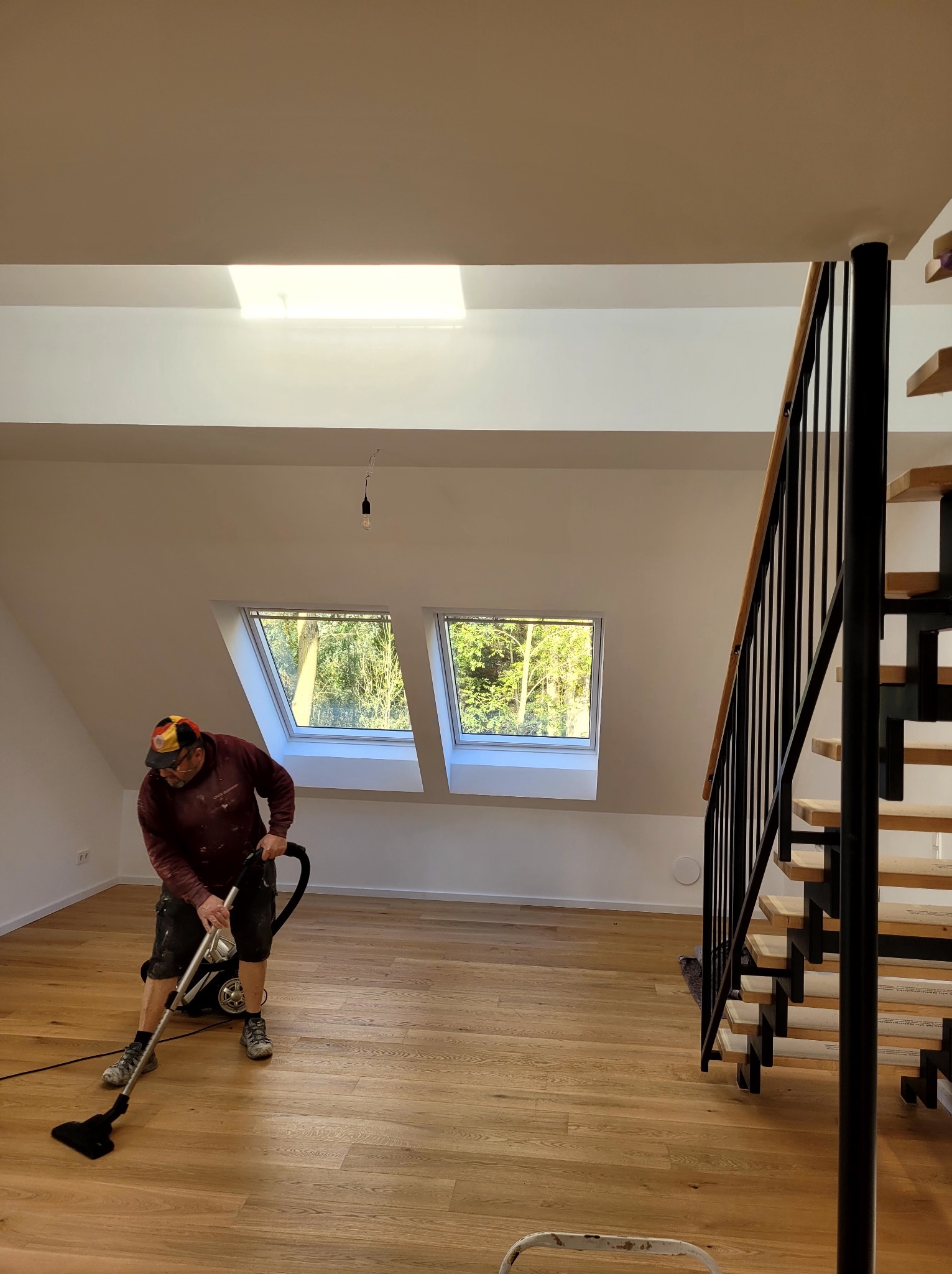 Remont poddasza: mężczyzna odkurza drewnianą podłogę po malowaniu ścian na biało. Widoczne okna dachowe i schody z czarną balustradą.