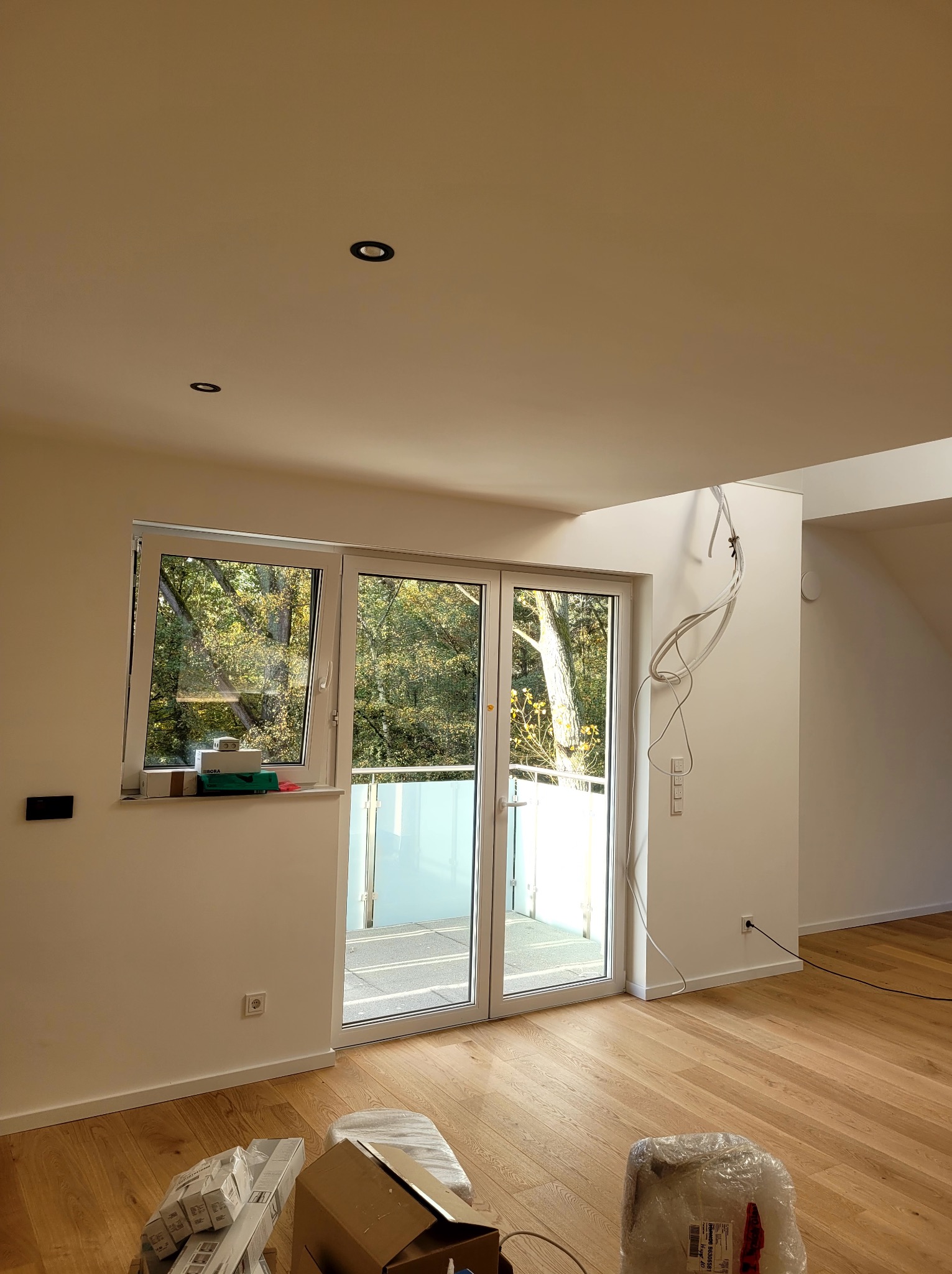 Jasne wnętrze po malowaniu: parkiet, białe ściany z gładzią, okno balkonowe z widokiem na zieleń, widoczne kable elektryczne i puszki farb. Minimalistyczny design.