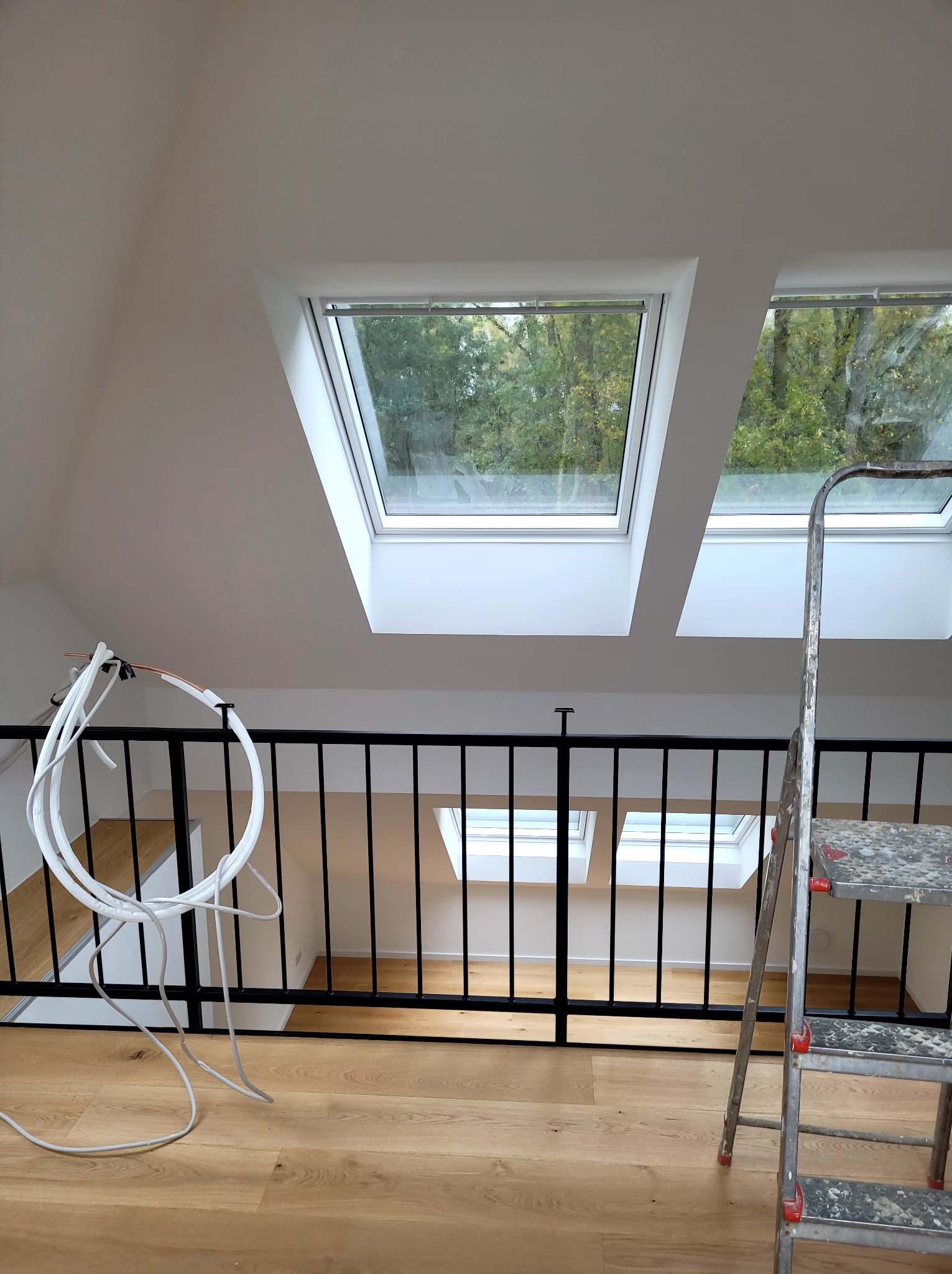 Poddasze po remoncie z dwoma oknami dachowymi, widok z antresoli z czarną balustradą. Na pierwszym planie drabina malarska i przewody. Ściany i sufit pomalowane na biało.