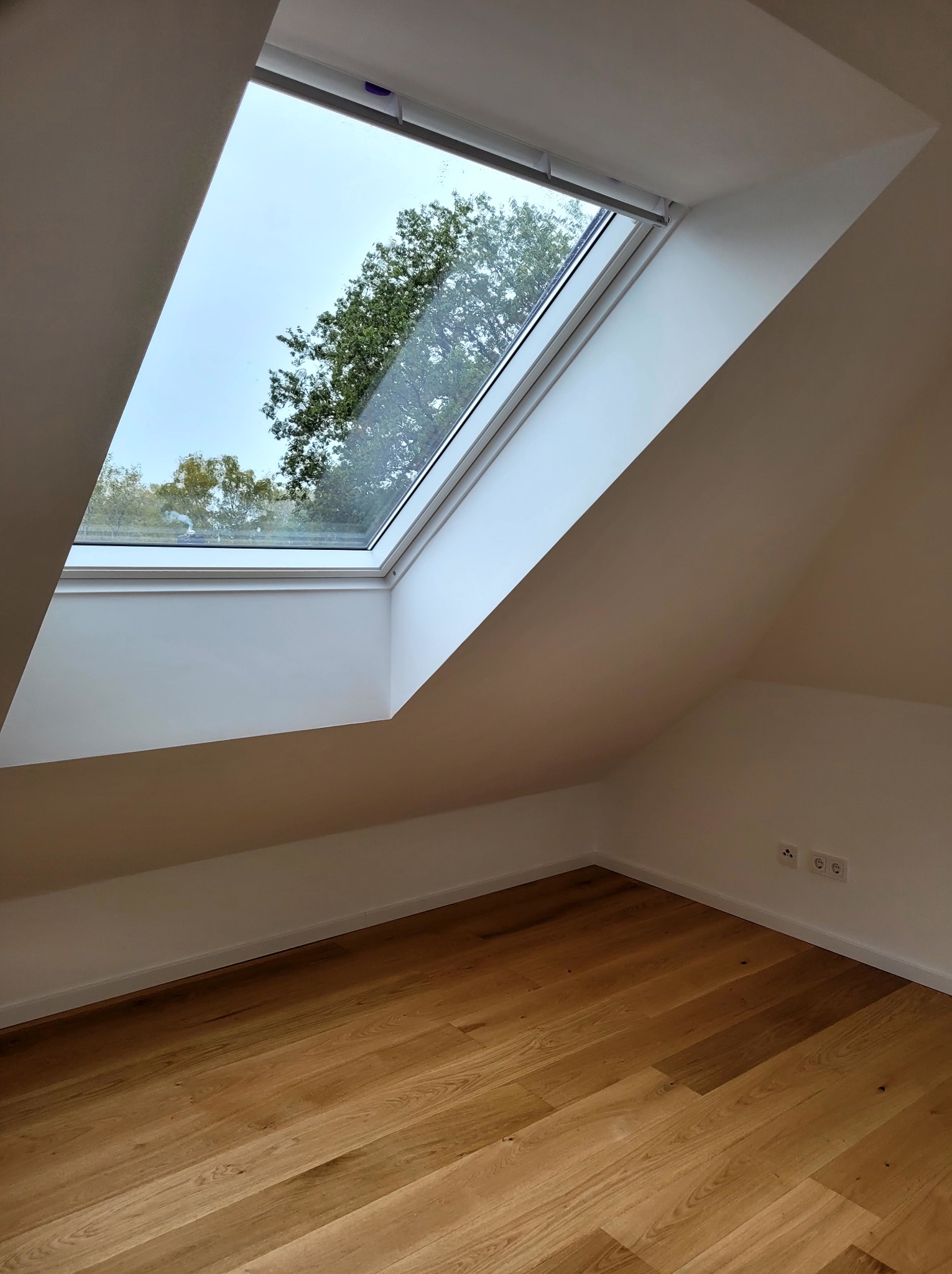 Jasne poddasze z oknem dachowym i drewnianą podłogą. Ściany pomalowane na biało, widoczne gniazdka elektryczne. Minimalistyczny design.