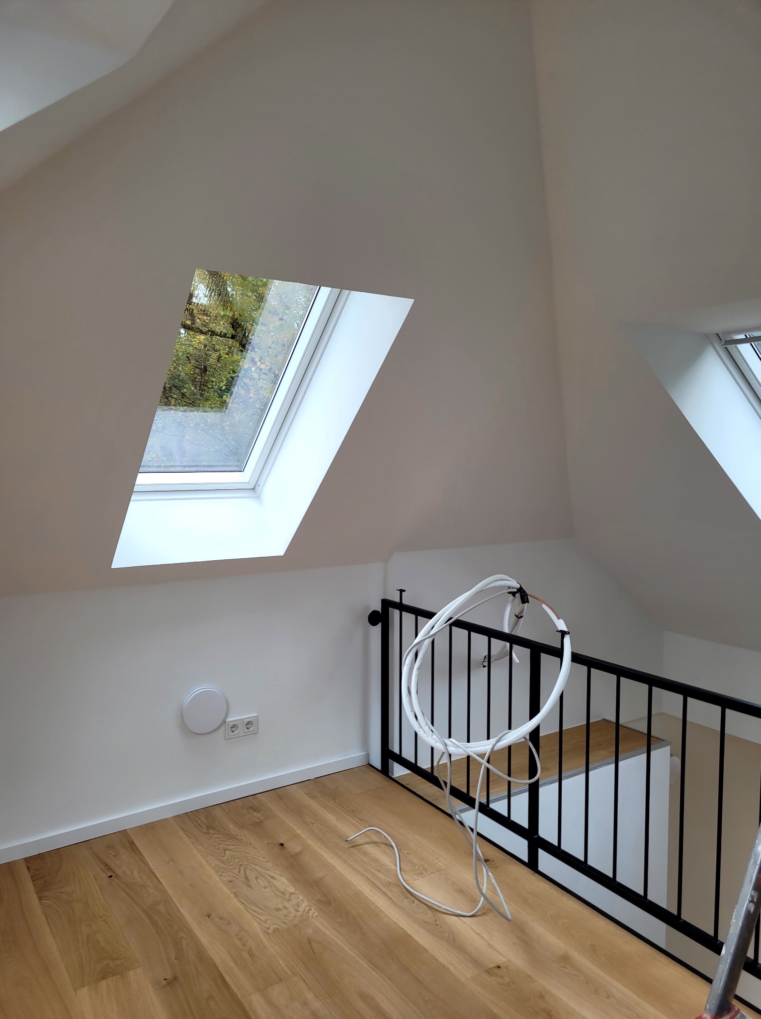 Białe poddasze po malowaniu z oknami dachowymi, drewnianą podłogą i czarną balustradą. Widoczne kable i gniazdka elektryczne. Jasne, naturalne światło.