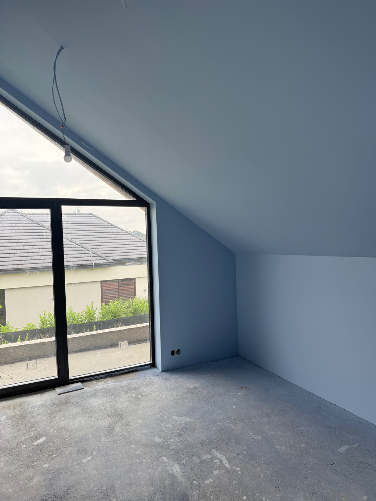 Poddasze po malowaniu na niebiesko, z dużym oknem i surową podłogą. Widoczny kabel z żarówką. Świeżo pomalowane ściany i sufit.