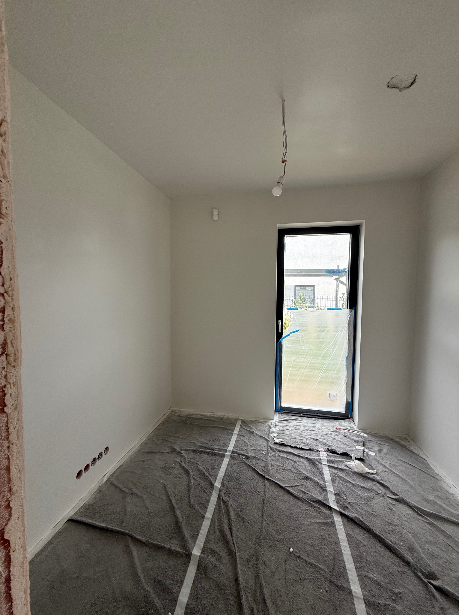 Wnętrze pokoju po malowaniu ścian na biało. Podłoga zabezpieczona folią ochronną. Widoczne gniazdka elektryczne i okno z widokiem na ogród.