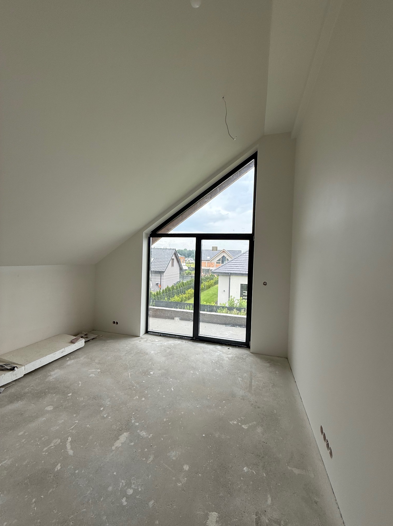 Jasne poddasze z oknem w kształcie trójkąta, świeżo pomalowane ściany i sufit na biało. Widoczne gniazdka elektryczne i surowa podłoga. Za oknem widok na domy.