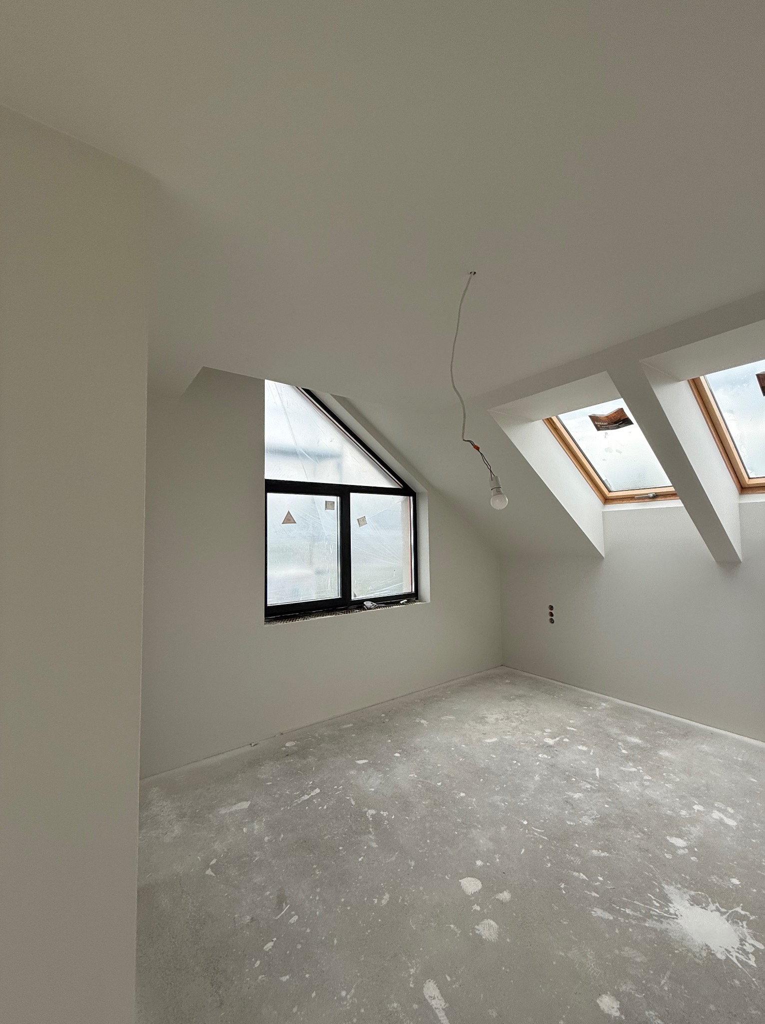 Wnętrze poddasza po gładziowaniu ścian i sufitu, z oknami dachowymi i trójkątnym oknem. Na podłodze widoczne ślady po pracach wykończeniowych.